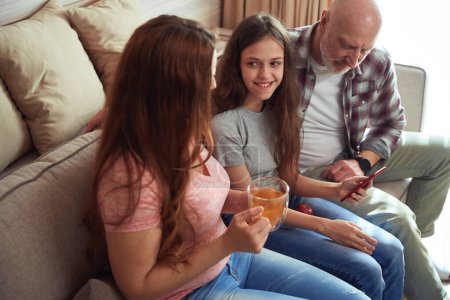 Téléchargez les photos : Adolescente agréable assise sur le canapé montrant quelque chose à sa mère et son père sur l'écran du smartphone - en image libre de droit