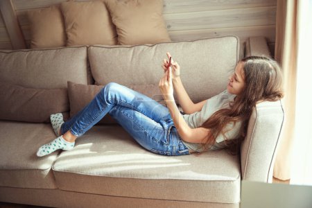 Téléchargez les photos : Joyeuse adolescente allongée sur un canapé tout en regardant quelque chose sur son téléphone portable dans ses mains - en image libre de droit