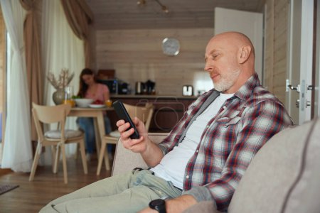 Photo for Focused male seated on sofa reading news on smartphone screen while his wife preparing food - Royalty Free Image