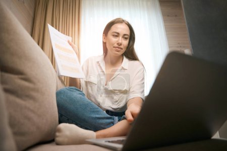 Photo for Beautiful woman holding papers and working on laptop remotely at home while sitting on the sofa - Royalty Free Image