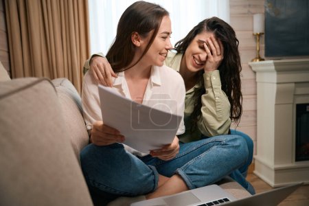 Foto de Señoras inteligentes sentadas en un cómodo sofá en la sala de estar, utilizando un ordenador portátil que trabaja de forma remota desde casa, comprobando, analizando documentos - Imagen libre de derechos