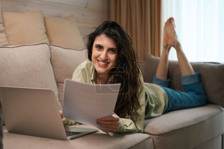 Téléchargez les photos : Happy femelle détenant des documents et travaillant sur ordinateur portable à distance à la maison tout en étant couché sur le canapé - en image libre de droit