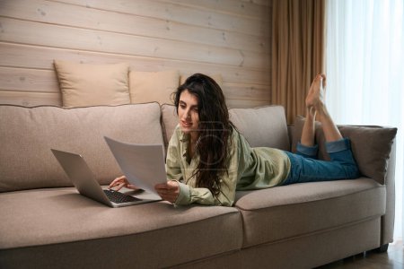 Téléchargez les photos : Jolie femme tenant des documents et travaillant sur un ordinateur portable à distance à la maison tout en étant couché sur le canapé - en image libre de droit