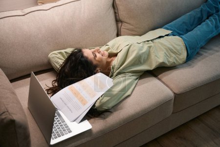 Téléchargez les photos : Femme heureuse ayant pause de travailler sur ordinateur portable à distance et la paperasserie tout en étant allongé sur le canapé et rêvant de vacances - en image libre de droit