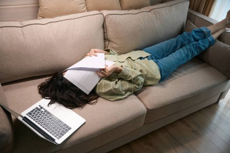 Foto de Mujer joven cansada trabajando en el ordenador portátil, mientras que la colocación en el sofá y ocultar la cara en las palmas de las manos sensación de dolor de cabeza migraña - Imagen libre de derechos