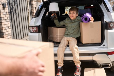 Téléchargez les photos : Le petit garçon est assis dans le coffre d'une voiture pendant que ses parents portent des choses dans la maison. - en image libre de droit