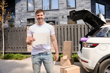 Téléchargez les photos : Jeune homme tient un morceau de papier dans ses mains et se tient sur le fond d'une voiture et une nouvelle maison. - en image libre de droit