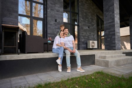 Téléchargez les photos : Jeunes parents et un enfant, embrassant, assis sur la terrasse d'une grande maison - en image libre de droit