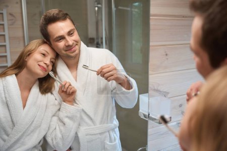 Téléchargez les photos : Souriant homme et femme brosser les dents dans la salle de bain, regarder dans le miroir - en image libre de droit