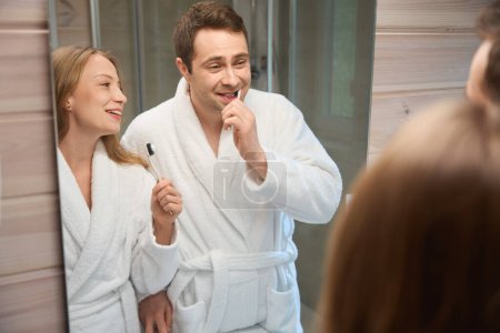 Téléchargez les photos : Heureux homme et femme se brossant les dents en s'émerveillant dans le miroir de la salle de bain - en image libre de droit