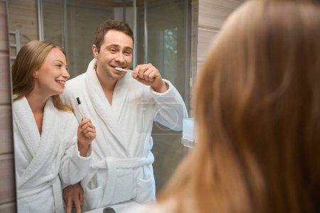 Photo for Happy man and woman in bathrobe brushing their teeth looking in the mirror - Royalty Free Image