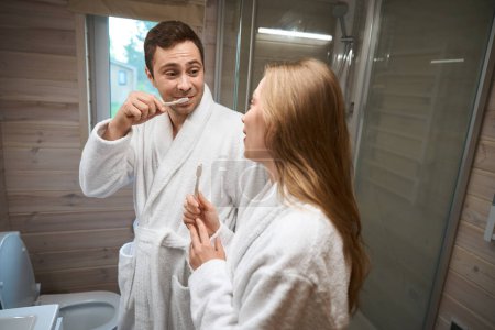 Foto de Hombre cepillándose los dientes y mirando a la mujer mientras está de pie en el baño - Imagen libre de derechos