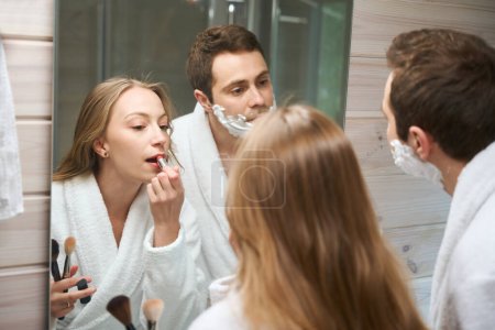 Photo for Young couple in the bathroom, woman paints her lips, man looks in the mirror - Royalty Free Image