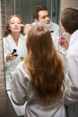Photo for Man shaving his beard woman putting on make-up while standing in the bathroom - Royalty Free Image