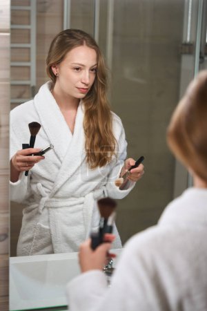 Téléchargez les photos : Jolie femme se regarde dans le miroir dans la salle de bain et tient des pinceaux blush - en image libre de droit