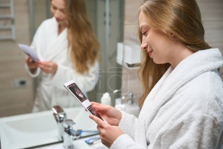 Téléchargez les photos : Femme enceinte dans la salle de bain regardant une photo de l'échographie - en image libre de droit