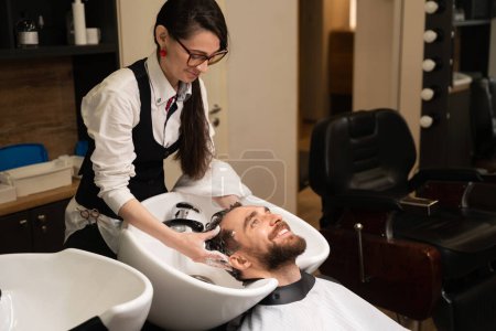 Téléchargez les photos : Jeune femme coiffeur lave et masse la tête d'un homme, le client est situé à un évier spécial - en image libre de droit