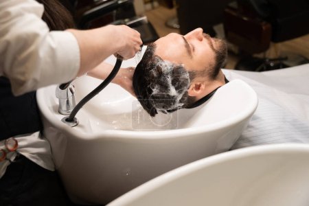 Téléchargez les photos : Barbier femme dans un salon lave un homme cheveux, un artisan utilise un shampooing spécial - en image libre de droit