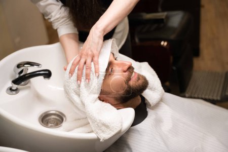 Foto de Peluquero hembra seca y masajea la cabeza de un hombre con una toalla suave, el cliente se encuentra en el fregadero especial - Imagen libre de derechos