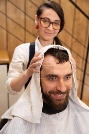 Téléchargez les photos : Mignon coiffeur sèche les cheveux du client avec une serviette douce, le mâle est situé dans la chaise de coiffure - en image libre de droit