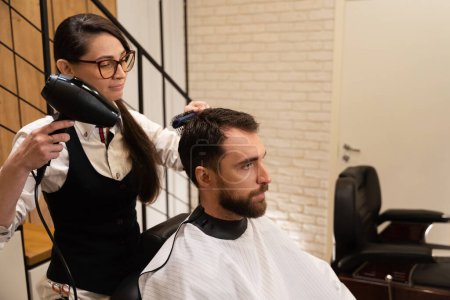 Téléchargez les photos : Coiffeur sèche les cheveux du client avec un sèche-cheveux professionnel, le mâle est situé dans la chaise de coiffure - en image libre de droit