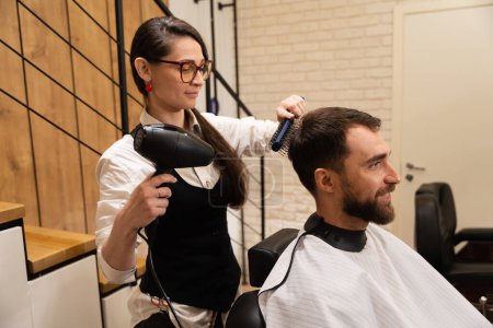 Téléchargez les photos : Femme coiffeur coiffeur clients cheveux avec un sèche-cheveux professionnel et brosse à coiffer, mâle assis dans une chaise de coiffeur - en image libre de droit