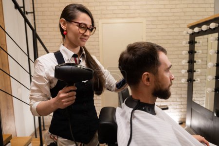 Téléchargez les photos : Jeune coiffeuse coiffant un client, elle utilise un sèche-cheveux et une brosse à coiffer - en image libre de droit