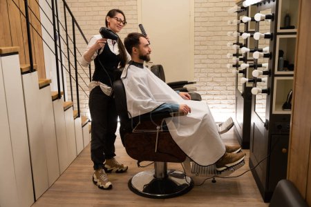 Téléchargez les photos : Jeune coiffeur femme coiffage clients cheveux avec sèche-cheveux et brosse à coiffer, homme assis dans la chaise de coiffeur - en image libre de droit