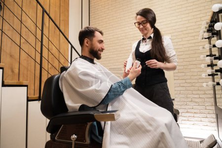 Téléchargez les photos : Femme dans un salon de coiffure démontre des produits de soins de la peau à un client, mâle est assis dans une chaise confortable - en image libre de droit