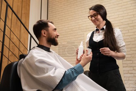 Téléchargez les photos : Jeune femme dans un salon de coiffure démontre des produits de soins masculins, le client est assis dans une chaise confortable - en image libre de droit
