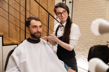 Téléchargez les photos : Barber femelle travaille avec une cliente dans un salon de coiffure moderne, dans son travail, elle utilise un produit coiffant - en image libre de droit