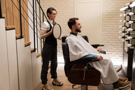 Photo for Craftswoman barber advises a client in a barbershop, she has a round mirror in her hands - Royalty Free Image