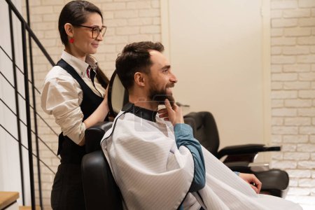 Photo for Young barber consults a man in a barbershop, she has a round mirror in her hands - Royalty Free Image