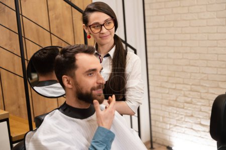 Téléchargez les photos : Jeune coiffeur montre sa coiffure du dos au client dans le miroir, le client sourit - en image libre de droit