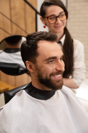 Téléchargez les photos : Barbier femme démontre dans le miroir au client sa coiffure du dos, l'homme sourit - en image libre de droit