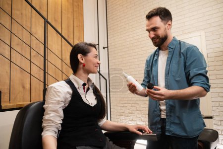 Téléchargez les photos : L'homme montre des pots de produits de soins à une jeune femme, les gens sont dans un coiffeur - en image libre de droit