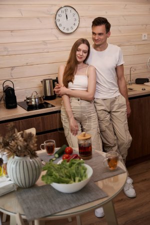 Téléchargez les photos : Jeune couple en vêtements décontractés debout dans la cuisine d'une maison de campagne dans une étreinte - en image libre de droit