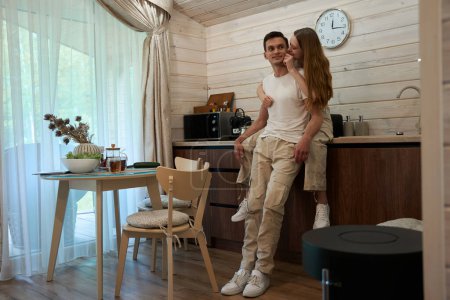 Téléchargez les photos : Magnifique femme en pyjama étreint son mari heureux tout en étant assis sur un comptoir de cuisine, les deux sourient - en image libre de droit