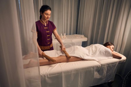 Téléchargez les photos : Thérapeute spa concentré roulant une paire de cannes de bambou sur la jambe nue du patient pendant la séance de massothérapie créole - en image libre de droit