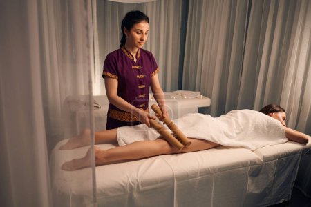 Foto de Serio masajista balanceo par de cañas de bambú sobre la parte posterior del muslo cliente femenino - Imagen libre de derechos