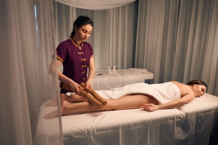 Téléchargez les photos : Femme cliente couchée couchée tandis que masseuse roulant paire de cannes de bambou sur ses muscles de mollet - en image libre de droit