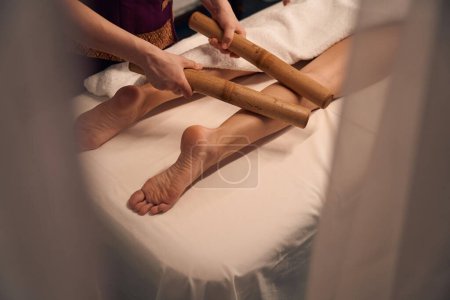Foto de Foto recortada del terapeuta de spa balanceo par de cañas de bambú sobre los músculos de la pantorrilla cliente - Imagen libre de derechos