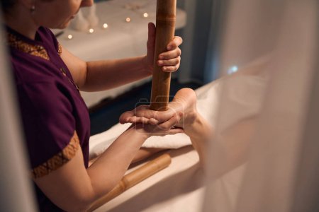 Téléchargez les photos : Photo recadrée de masseuse pressant avec extrémité de canne en bambou sur le point réflexe du pied du client - en image libre de droit