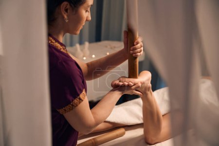 Téléchargez les photos : Photo recadrée de masseuse stimulante avec pointe réflexe d'extrémité de canne de bambou sur le pied patient féminin - en image libre de droit