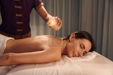 Photo for Young woman lying prone with eyes closed while masseuse pouring melted wax on her naked back - Royalty Free Image