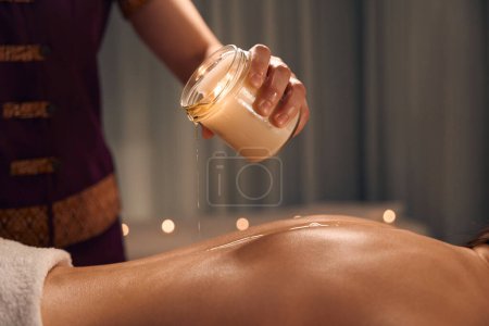 Photo for Cropped photo of massotherapist pouring melted wax from jar on female back - Royalty Free Image