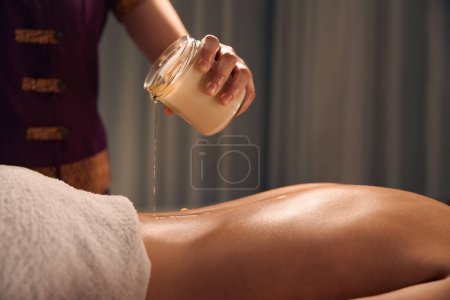 Photo for Cropped photo of spa therapist pouring melted wax from jar on female patient back - Royalty Free Image