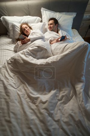 Téléchargez les photos : Homme et femme en peignoirs chauds flirtant couchés au lit et bavardant sur les smartphones les uns avec les autres, les gens oubliant la vraie vie à cause d'Internet - en image libre de droit