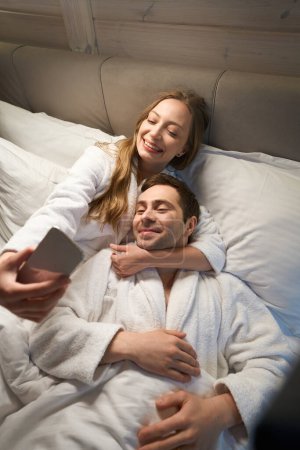 Foto de Hermosa mujer abrazando a su apuesto marido por el cuello haciendo selfie en el teléfono inteligente, pareja descansando y divirtiéndose tumbada en la cama en cálidos albornoces suaves - Imagen libre de derechos