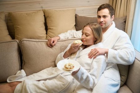 Photo for Beautiful woman in comfortable bathrobe enjoying taste of fresh sweet dessert leaning on her husband, couple resting and relaxing sitting on sofa in vacation home - Royalty Free Image